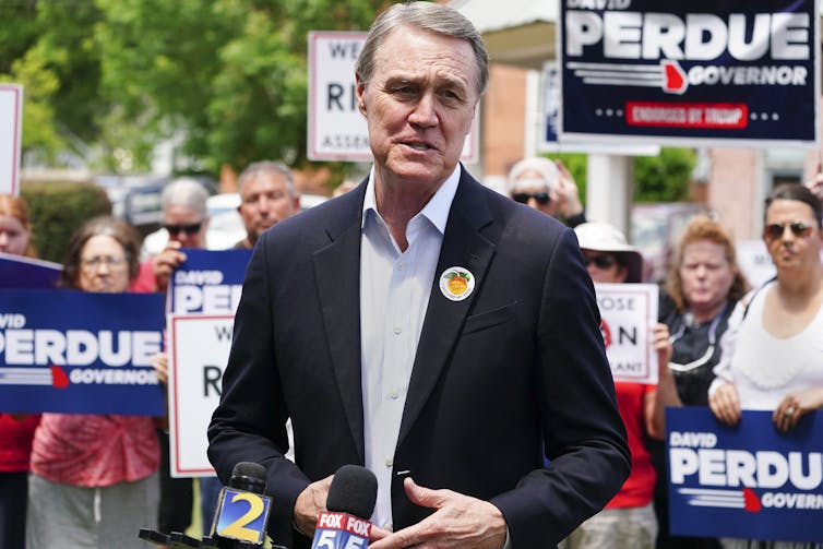 A man with gray hair wearing a blazer at a campaign rally with signs held behind him.