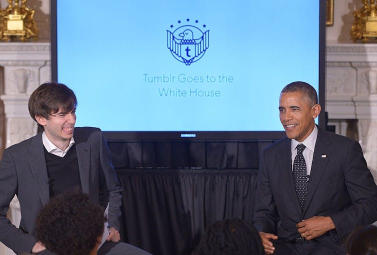 Two smiling men sit in front of a screen.