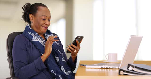 Une femme souriante regarde son cellulaire, assise devant un ordinateur