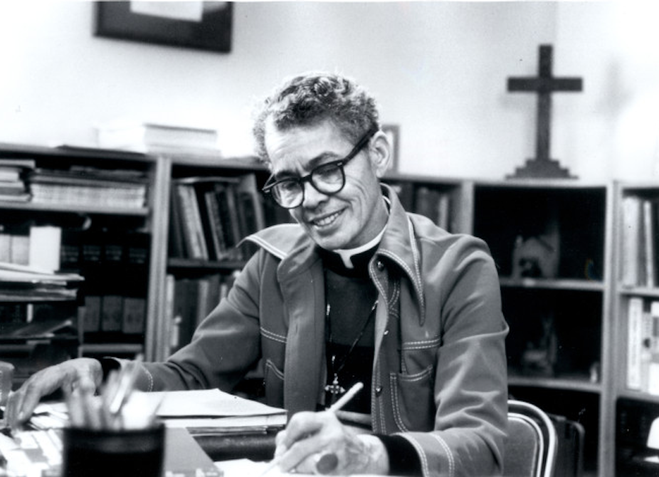 A person with short hair and glasses sits and smiles while she reads at a table, with a cross on the bookshelf behind.