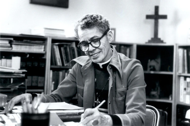A person with short hair and glasses sits and smiles while she reads at a table, with a cross on the bookshelf behind.