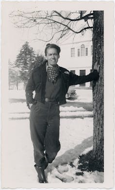 A young woman in thick pants, jacket and scarf leans against a tree on a snowy day.