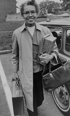 A middle-aged woman holding a briefcase and purse and wearing a raincoat walks between cars in a parking lot.