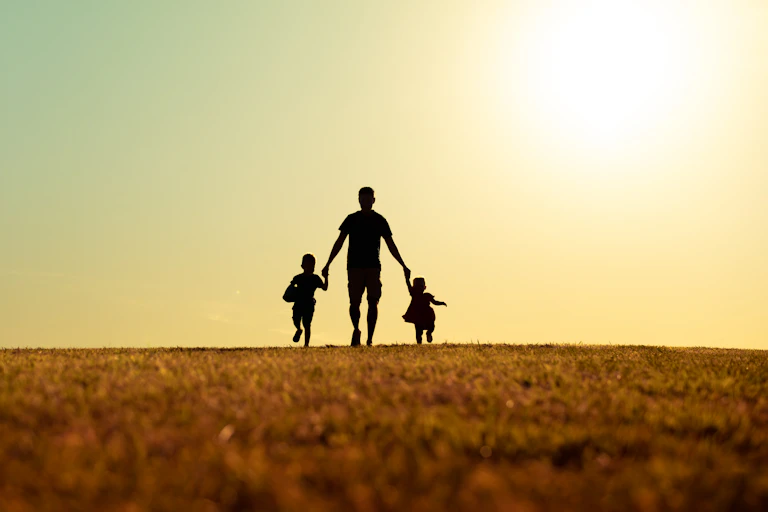 Silhouette of a father walking with son and daughter in park at sunset.