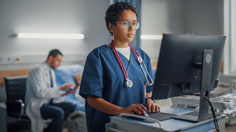 Nurse standing in front of a computer while a doctor sits beside a patient.