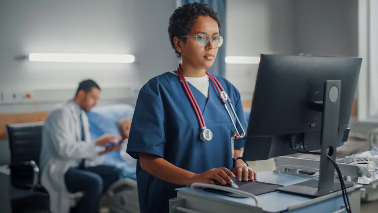 Nurse standing in front of a computer while a doctor sits beside a patient.