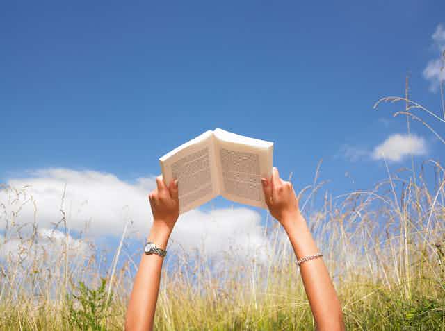 A young person's arms are extended toward a blue sky with several white clouds as the person grasps an open book that they are reading while laying in a tall field of grass.