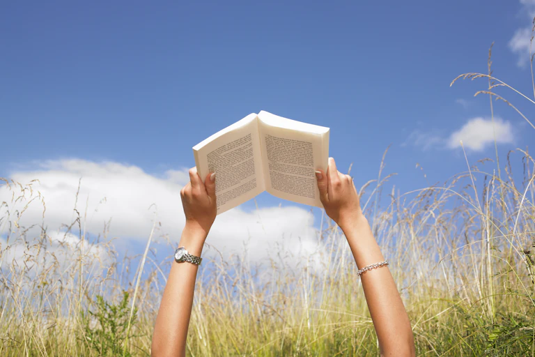 A young person's arms are extended toward a blue sky with several white clouds as the person grasps an open book that they are reading while laying in a tall field of grass.