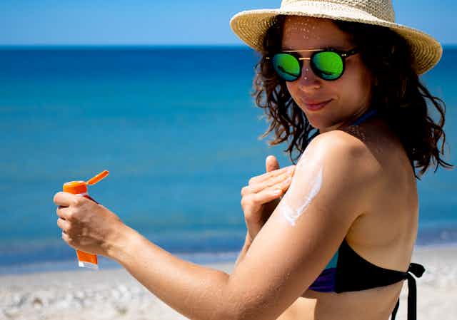 Une jeune femme brune à peau claire se protège du soleil avec un chapeau, des lunettes et se met de la crème solaire