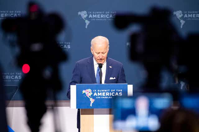 Joe Biden stands at a podium at the Summit of the Americas with silhouettes of TV cameras in the foreground.