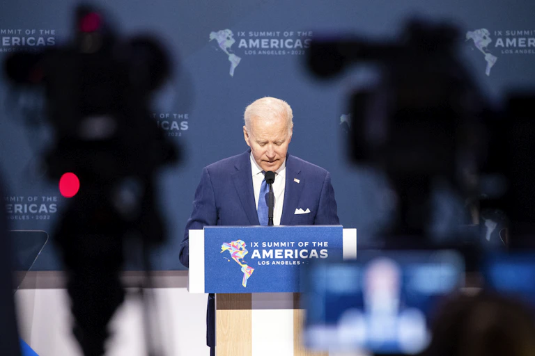Joe Biden stands at a podium at the Summit of the Americas with silhouettes of TV cameras in the foreground.
