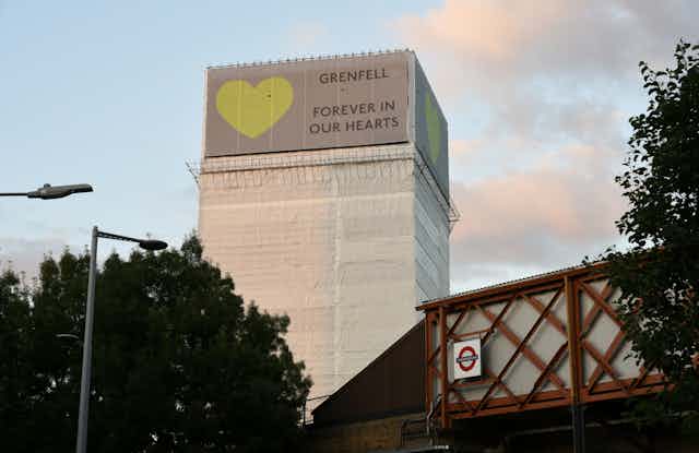 A high-rise building wrapped in white plastic sheets with a green heart and the message, Grenfell forever in our hearts.