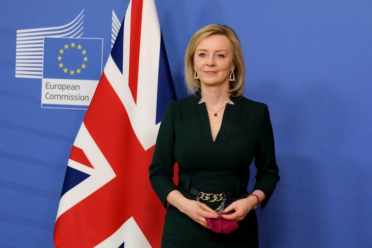 Liz Truss stands in front of a UK flag, and a background that has the European Commission logo.