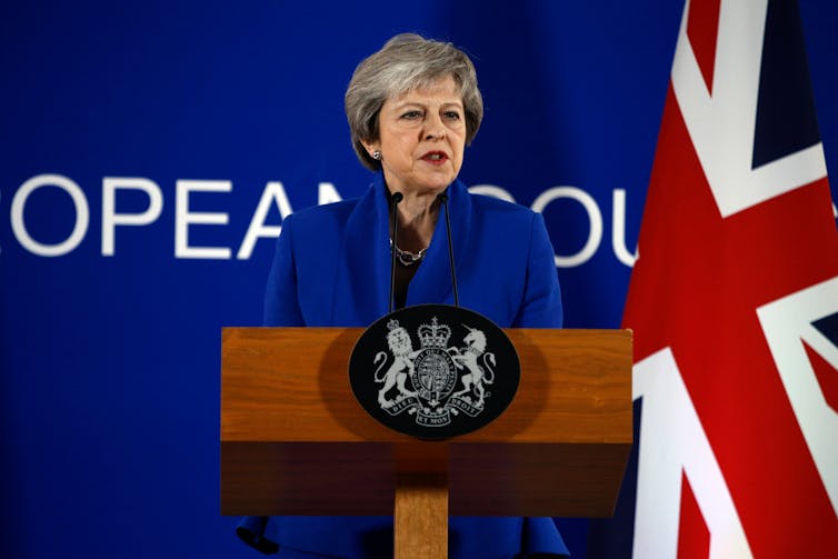 Theresa May speaking at a podium in front of a UK flag and blue background