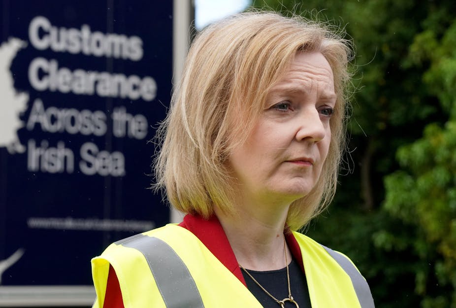 UK foreign secretary Liz Truss looks forlorn in a high-vis vest. Behind her is a billboard reading Customs Clearance Across the Irish Sea.