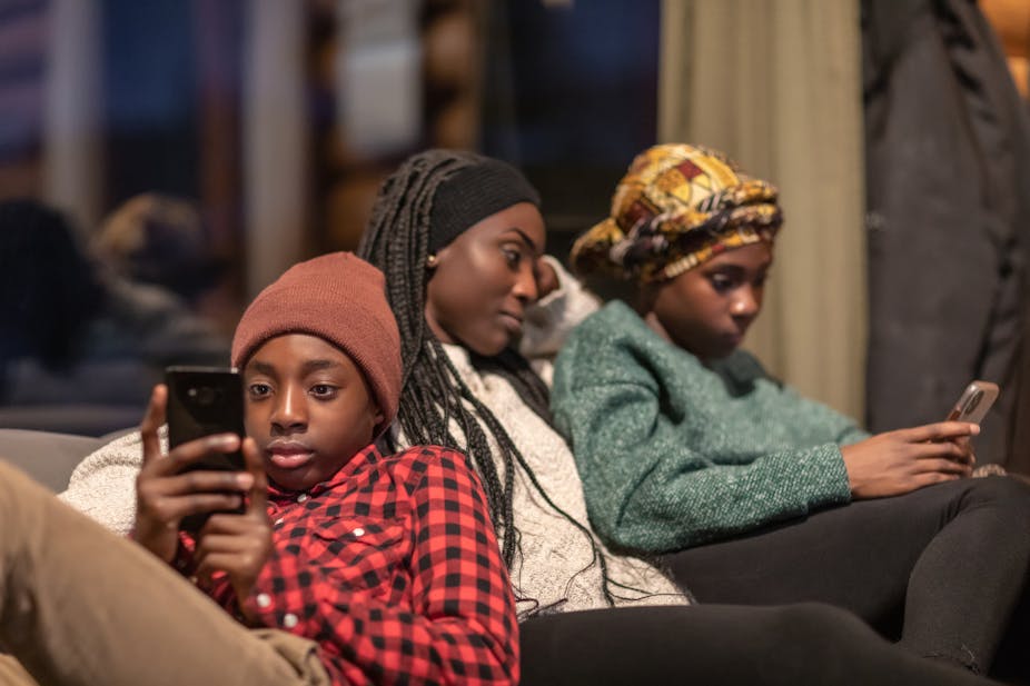 An adolescent boy leans against his mother as he holds his smart phone and engages with it. His mother is cuddled up to his sister and they both look at her smart phone.