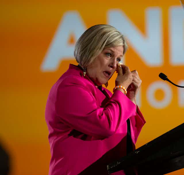 A blonde woman in a pink jacket wipes away a tear.