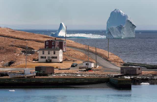 A point of land with a few buildings and boats with a very large and elongated iceberg in the water behind it. 