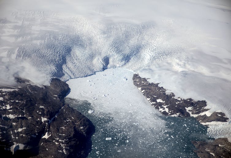 An aerial view of a glacier with large and small chunks of ice floating in the water at its foot, with rocky slopes on either side of the narrow fjord.