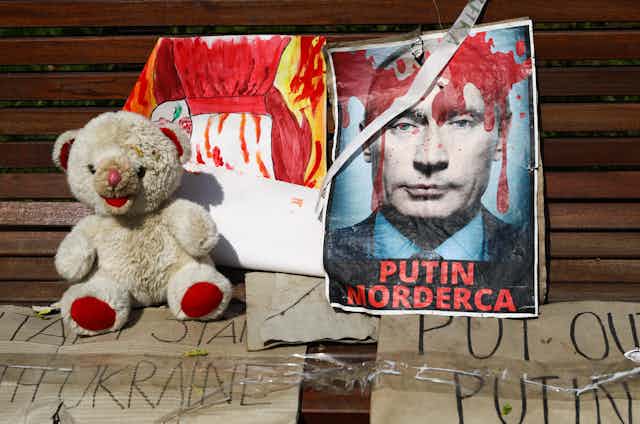 A teddy bear sits next to a headshot of Putin, which is covered in fake blood and says 'Putin, murderer.'
