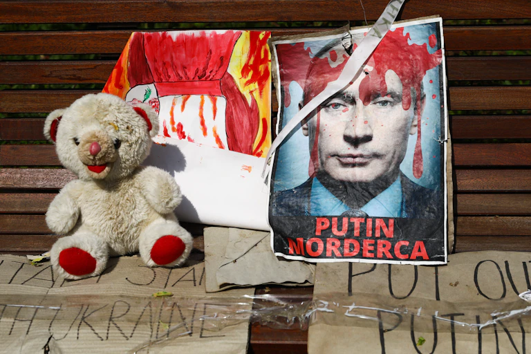 A teddy bear sits next to a headshot of Putin, which is covered in fake blood and says 'Putin, murderer.'