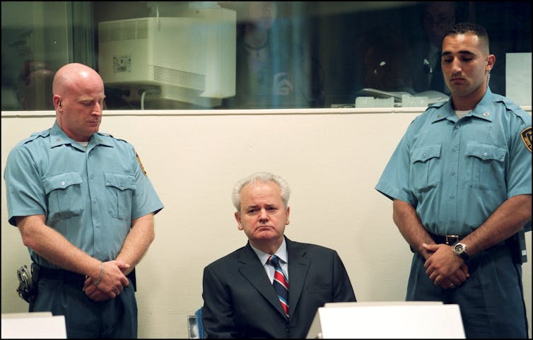 Milosevic sits in a suit, with two guards in blue shirts on either side of him.