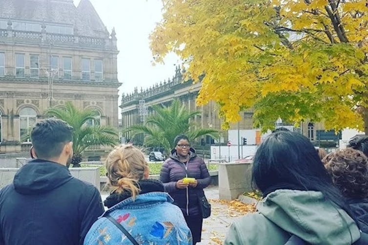 Tourists with their back to the camera listen to a tour guide in an outdoor setting.