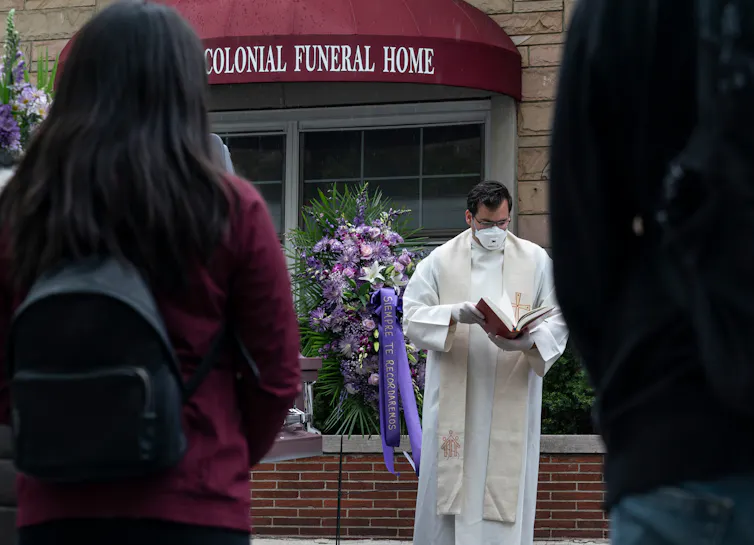 A priest stands outside a building wearing a mask