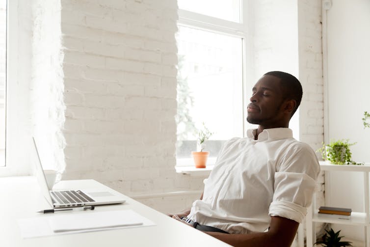 A man in a chair in white shirt, in a white room, with his eyes closed.