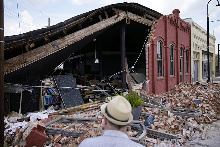 A man in a hat faces a building with a partially collapsed brick wall.