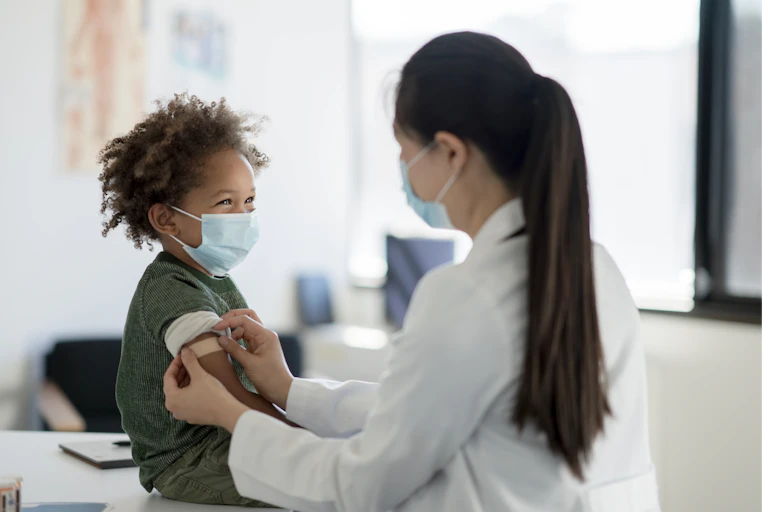 A smiling young boy wearing a mask sits on an exam table as his female doctor places a bandage on his arm after receiving a vaccine.