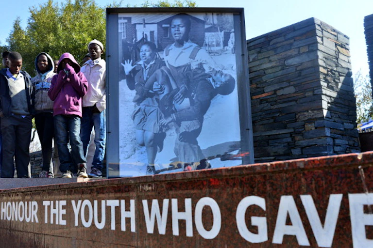A handful of boys stand next to a memorial showing a schoolboy, with blood oozing from his face, being carried by an older boy, whille the dead boy's sister walks beside him.