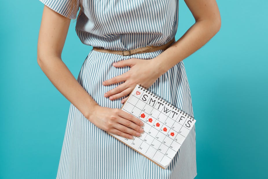 A woman holds her abdomen in pain, while clutching a calendar indicating when her period will arrive during the month.