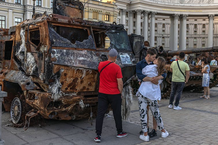 A young couple embraces in front of wrecked Russian military vehicles in central Kyiv as other people walk past.