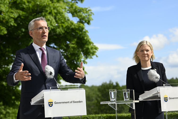 A middle-aged white man and white woman dressed in a business suits stand behind lecturns during a press conference.