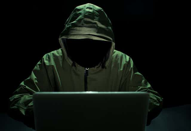A hooded faceless figure sits at a laptop.