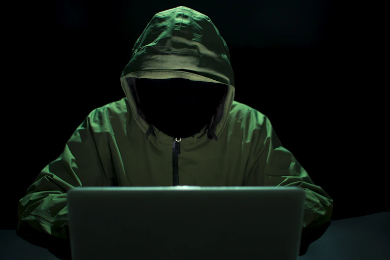 A hooded faceless figure sits at a laptop.