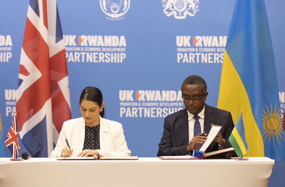 Priti Patel and Vincent Biruta sit at a table, each signing a document, in front of UK and Rwandan flags