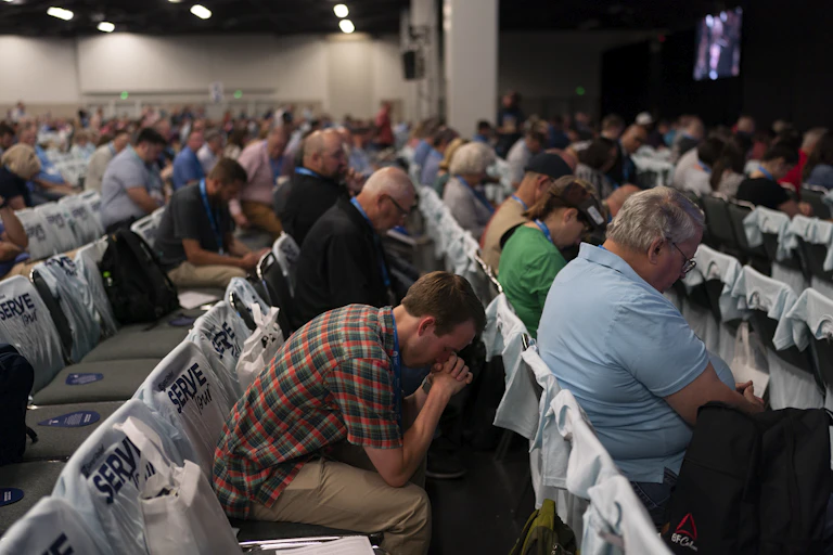 People sitting in neatly arranged rows pray with their heads bowed.