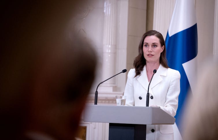 A white woman wearing a white blazer stands behind a lectern and talks to reporters.