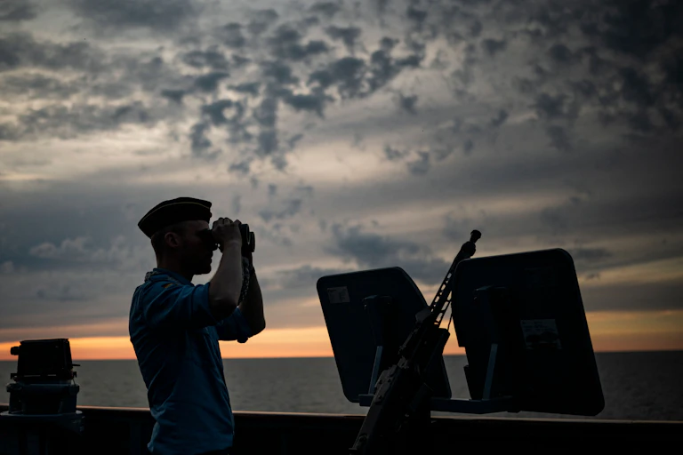 With the sun setting in the background, a silhouette of a soldier standing near an anti-aircraft weapon while looking through a pair of binoculars.