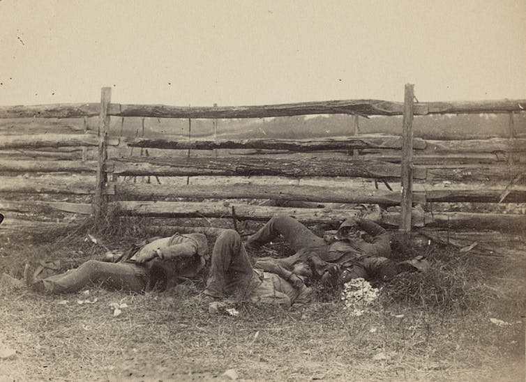 Une photo en noir et blanc de trois corps allongés à côté d'une clôture à rails fendus