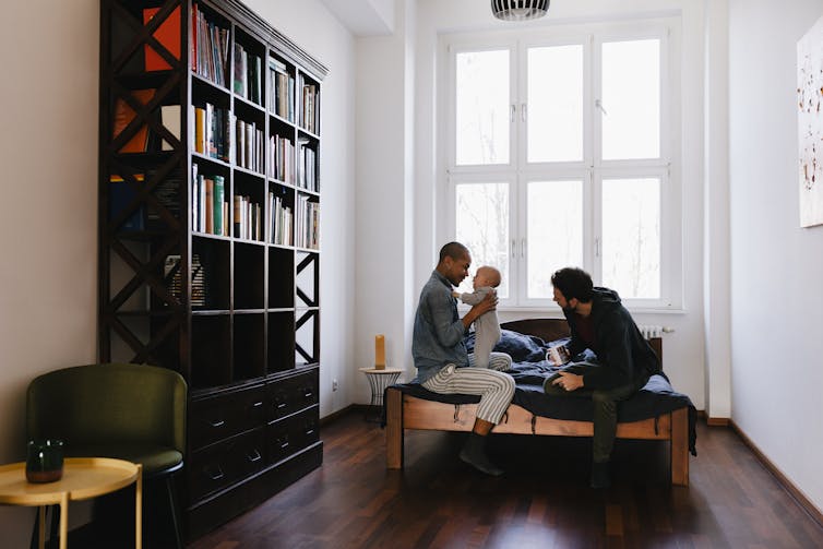 two men sit on bed with baby with a tall bookshelf against the wall