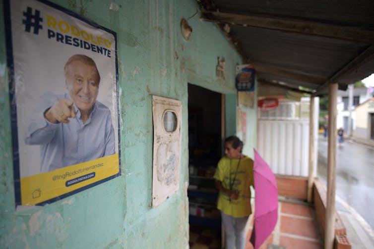 A woman holding a pink umbrella is seen near a poster poster promoting presidential candidate Rodolfo Hernandez.