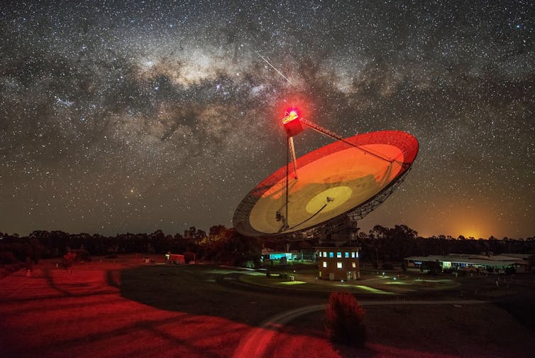 Nighttime photograph showing a large radio dish with a glowing red light with stars and the Milky Way visible in the background.