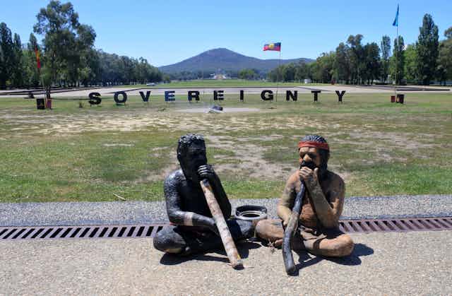 Two statues of First Nations people playing didgeridoos in front of a sign that reads 'sovereignty' besides Aboriginal and Torres Strait Islander Flags.