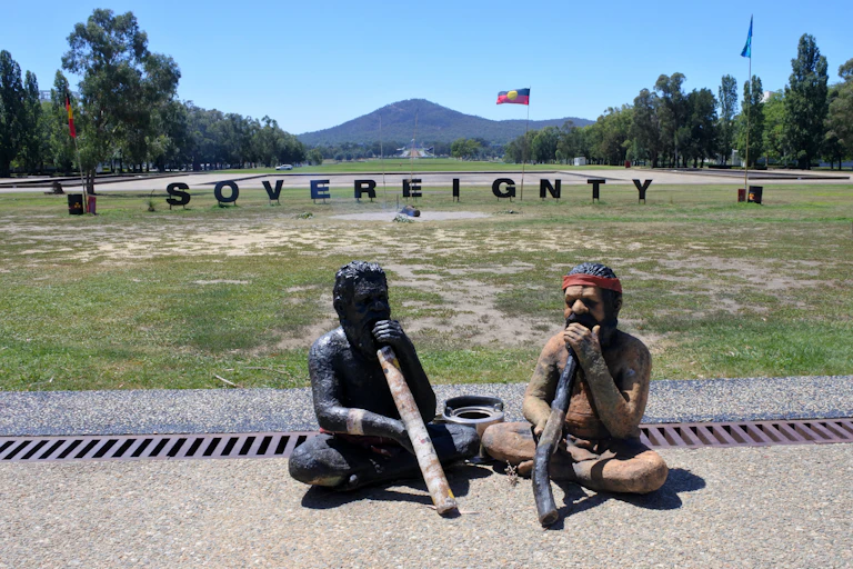 Two statues of First Nations people playing didgeridoos in front of a sign that reads 'sovereignty' besides Aboriginal and Torres Strait Islander Flags.