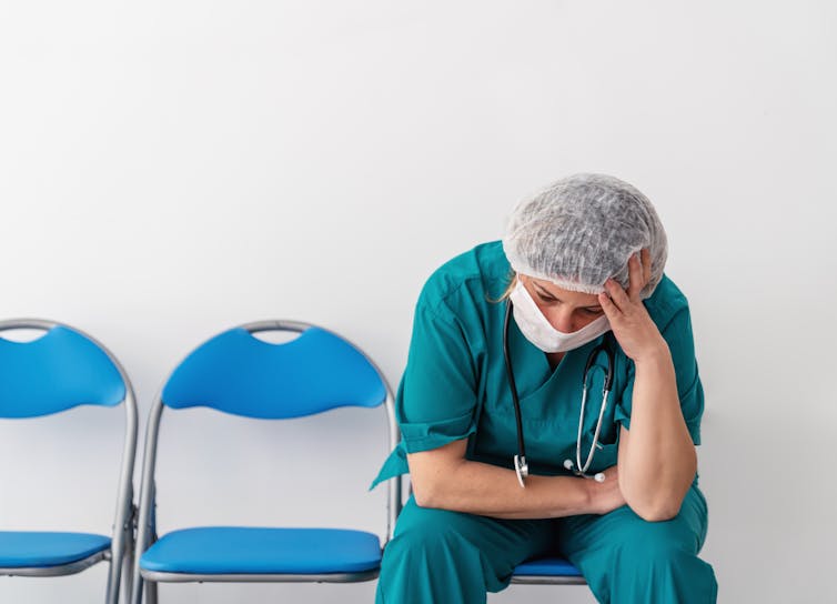 A person in scrubs, a face mask and a hair covering, with a stethoscope are their neck, sits alone in a row of folding chairs with head in hand