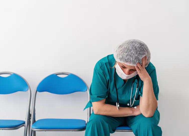 A person in scrubs, a face mask and a hair covering, with a stethoscope are their neck, sits alone in a row of folding chairs with head in hand