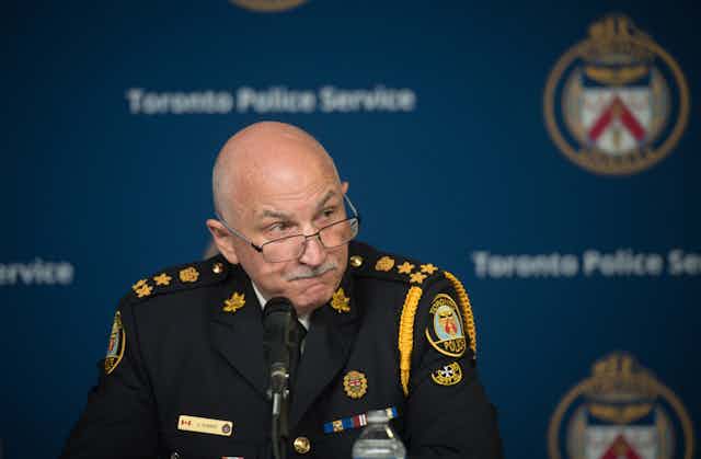 A bald man with glasses in uniform sits with a microphone in front of him. The words Toronto Police Service are on the wall behind him.